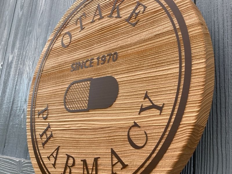 薬局ロゴの木製看板を斜めから捉えたクローズアップ。木の温かい木目と、こげ茶色の立体的な文字彫刻がわかる。