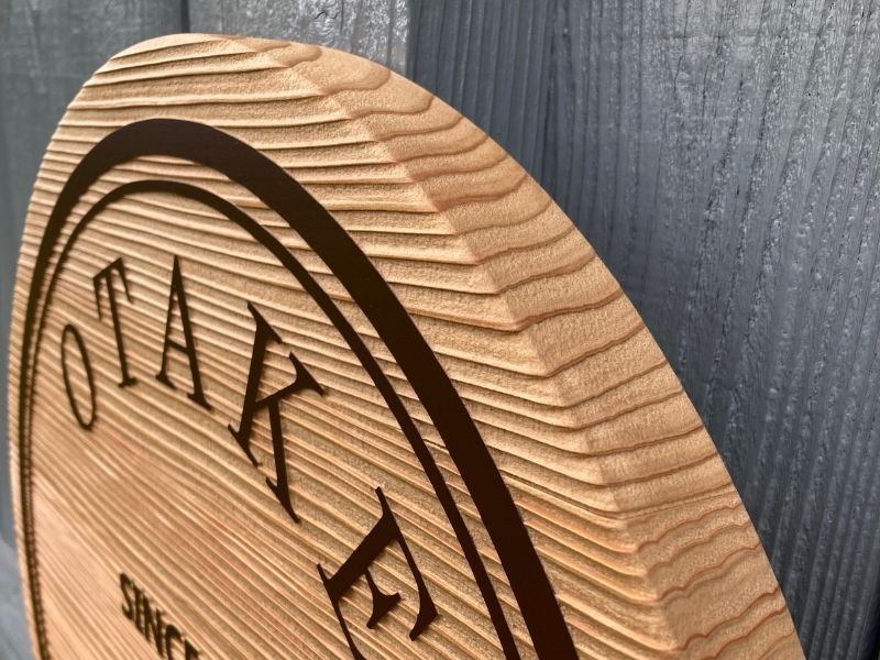 木製看板の縁（ふち）部分を真横からクローズアップした写真。丁寧に削り出された円形の木肌と、彫り込まれたラインの細部が写っている。