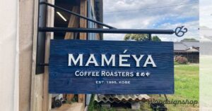 「MAMÉYA Coffee Roasters」の青い木製看板が建物外壁に取り付けられている。白い立体的な文字が映えるデザイン