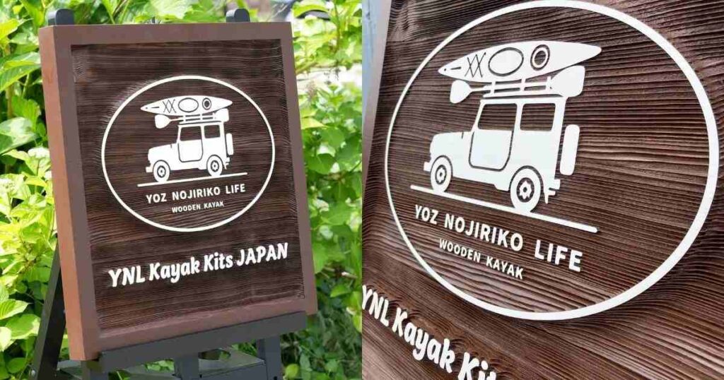 木製看板がイーゼルに立てかけられ、緑豊かな道沿いに設置されている様子。看板にはカヤックと車のロゴと「YOZ NOJIRIKO LIFE WOODEN KAYAK」の文字が描かれている。