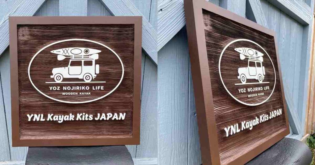 木製看板のクローズアップ。杉材に施されたサンドブラスト加工の浮き彫りが際立ち、車に載せられたカヤックと「YOZ NOJIRIKO LIFE WOODEN KAYAK」の文字が確認できる。
