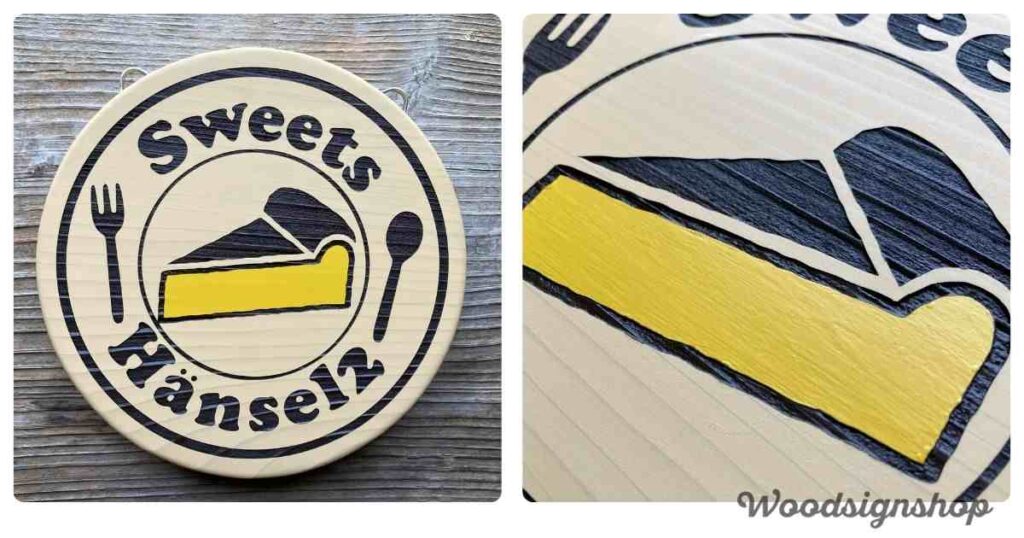 平らな木目の背景に置かれた、丸い木製看板の全体像。看板には「Sweets Hänsel2」の文字、黄色いチーズケーキのイラスト、フォークとスプーンが黒い文字と線で描かれている。両サイドには吊り下げ用の金具が見える。