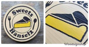 平らな木目の背景に置かれた、丸い木製看板の全体像。看板には「Sweets Hänsel2」の文字、黄色いチーズケーキのイラスト、フォークとスプーンが黒い文字と線で描かれている。両サイドには吊り下げ用の金具が見える。