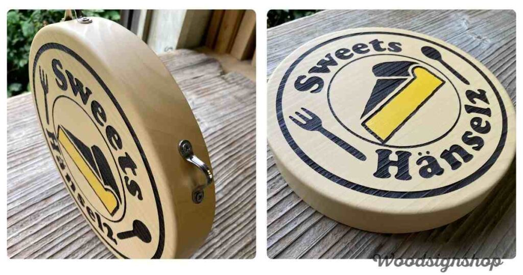 木目調の丸い木製看板が、屋外の木製ベンチに立てかけられている様子。看板には「Sweets Hänsel2」の文字と、チーズケーキ、フォーク、スプーンのイラストが描かれている。