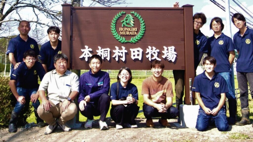 北海道・本桐村田牧場のスタッフとウッドサインショップ林が木製看板の前で記念撮影している様子。