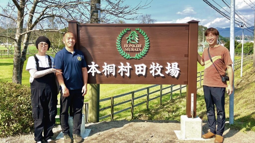 北海道本桐村田牧場の新しい木製看板の前で、牧場関係者と看板製作者の3名が記念撮影。看板には「HONKIRI MURATA 本桐村田牧場」の文字とロゴ。