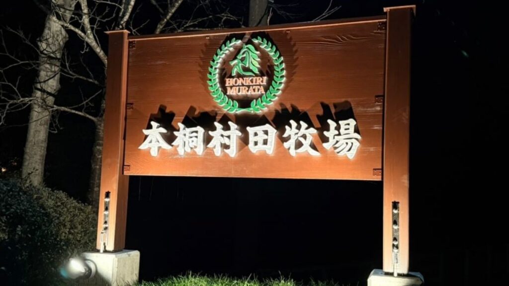 夜間のスポットライトで下から照らされた、本桐村田牧場の重厚な木製看板。暗闇の中で文字とロゴが鮮明に浮かび上がっている。