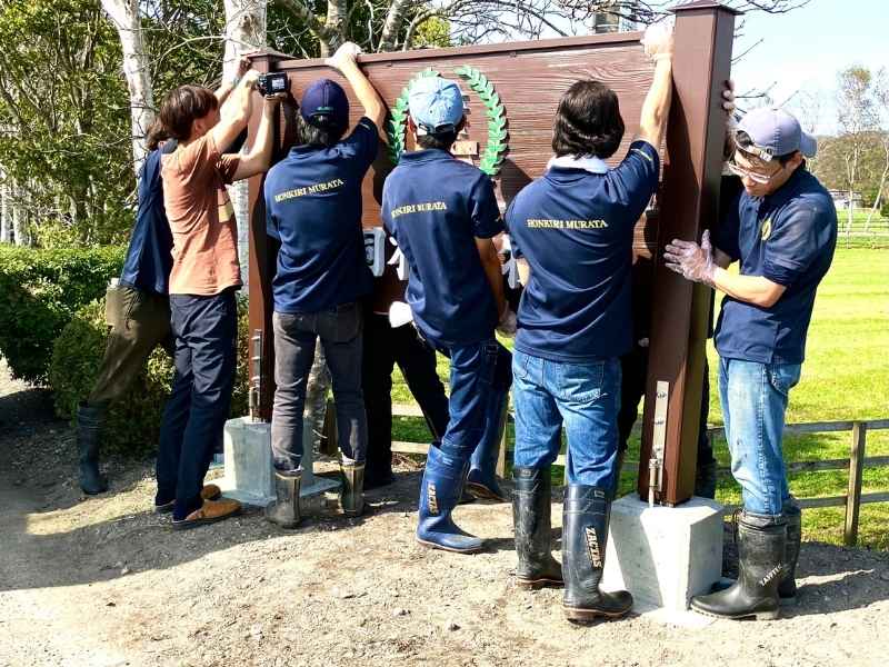 青空の下、本桐村田牧場の新しい木製看板を皆で協力して設置する瞬間。