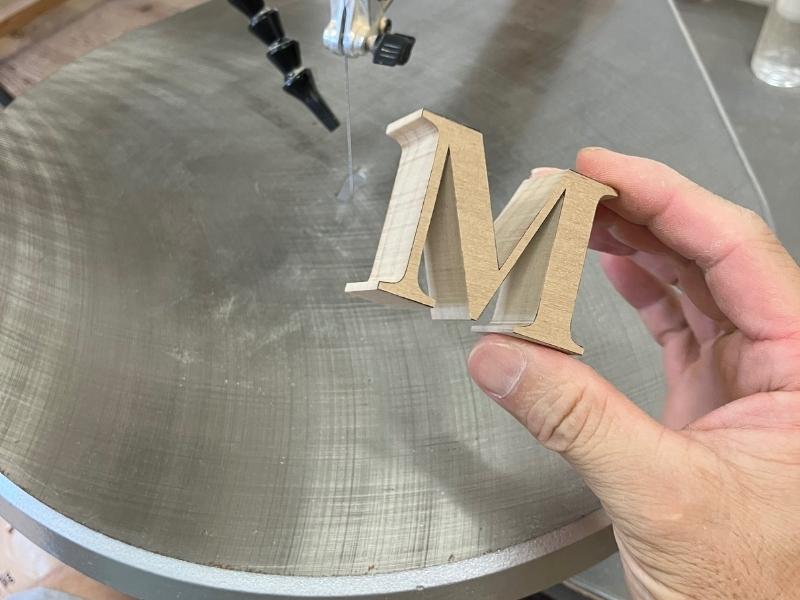 木製看板用のアルファベット「M」を糸鋸で切り出した直後の様子。手で持ち、文字の厚みと木の素材感がわかる。