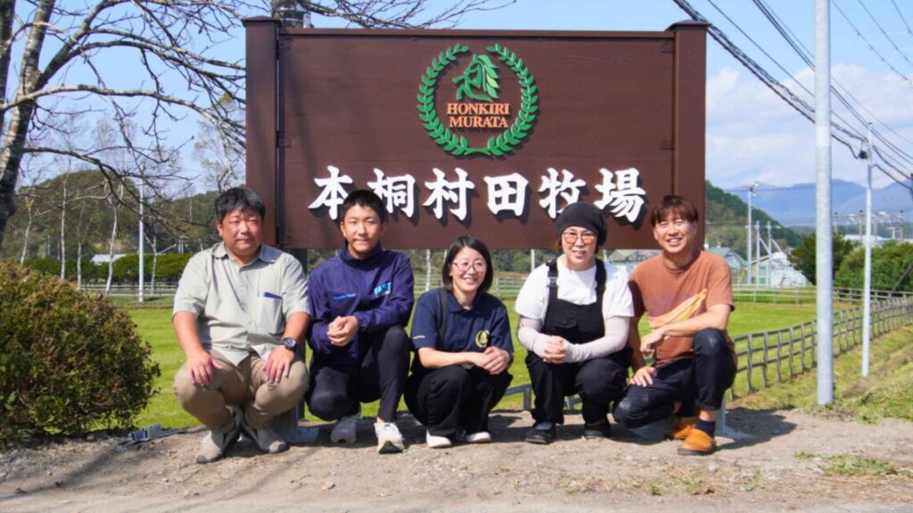 本桐村田牧場の看板前で撮影された、村田社長ご家族と私たちの集合写真。