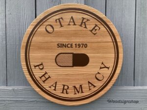 木製看板の全体像。OTAKE PHARMACYの円形ロゴとカプセルアイコン、SINCE 1970の文字が彫刻されている。
