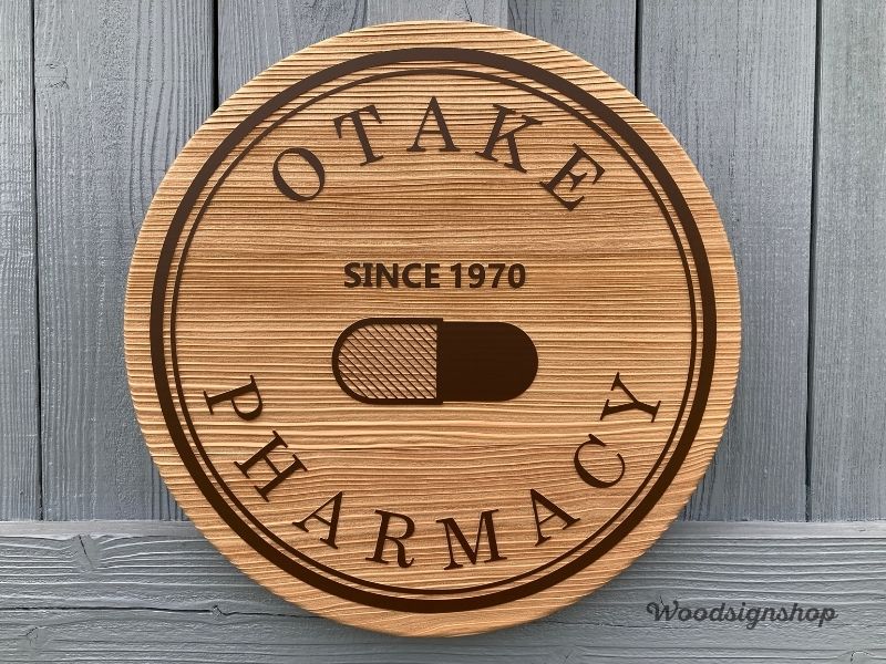 木製看板の全体像。OTAKE PHARMACYの円形ロゴとカプセルアイコン、SINCE 1970の文字が彫刻されている。