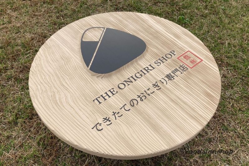 芝生の上に置かれた丸い木製看板。サンドブラスト加工の木目が目立たない角度だが、おにぎりマーク、店名、所在地はすべて明確に読み取れる。