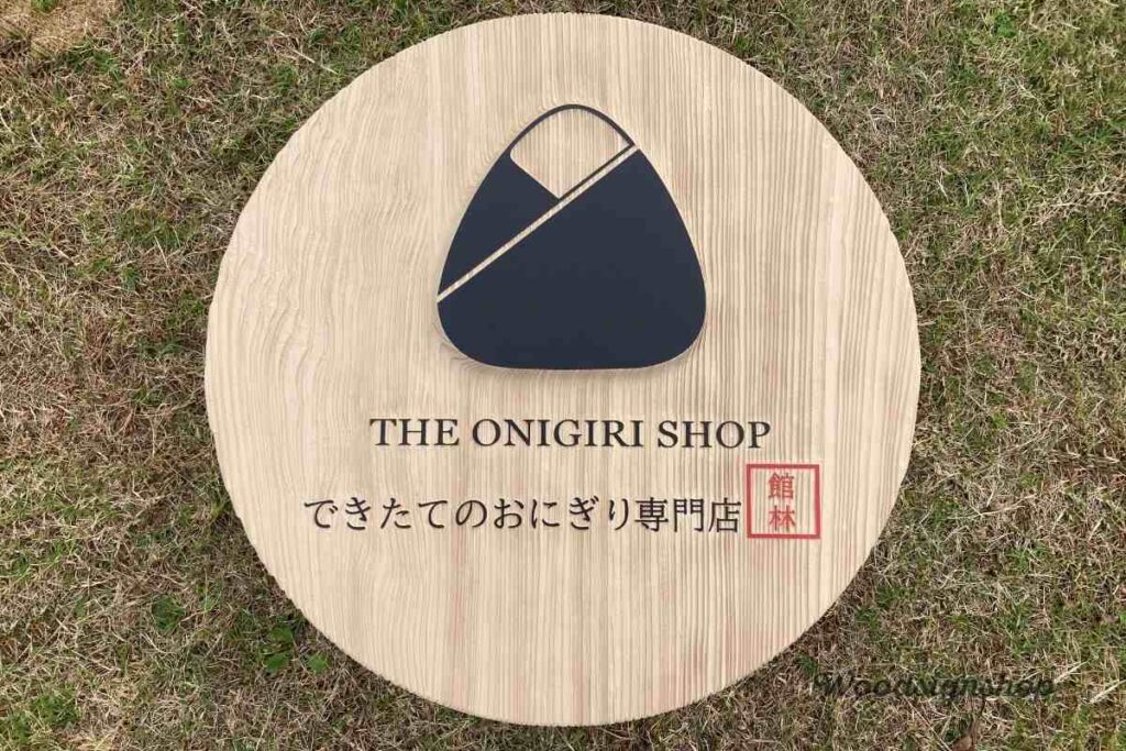 縦方向のサンドブラスト加工で木目を際立たせた、丸い木製看板。おにぎりマークが立体的な凸、文字が凹で彫刻され、強い日光で陰影が強調されている。群馬県館林市のおにぎり専門店用。