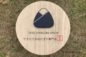 縦方向のサンドブラスト加工で木目を際立たせた、丸い木製看板。おにぎりマークが立体的な凸、文字が凹で彫刻され、強い日光で陰影が強調されている。群馬県館林市のおにぎり専門店用。