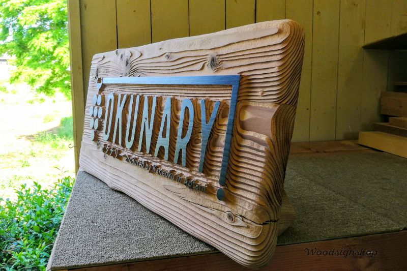 緩やかな曲線を描く木目に黒い文字で「OKUNARI」と刻まれた、無塗装の自然な風合いの木製看板