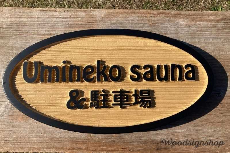 楕円形の木製看板に「Umineko sauna & 駐車場」と黒い文字で彫られた案内看板。