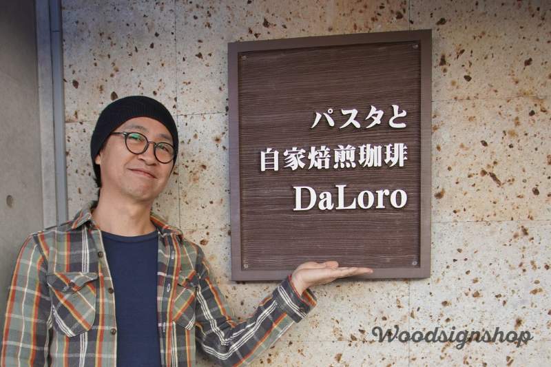 鹿児島中央駅近くの人気パスタ店DaLoro(ダローロ)の入り口に設置された、経年変化で風合いを増した茶色のオーダーメイド木製看板と、その横で微笑む制作者の男性。