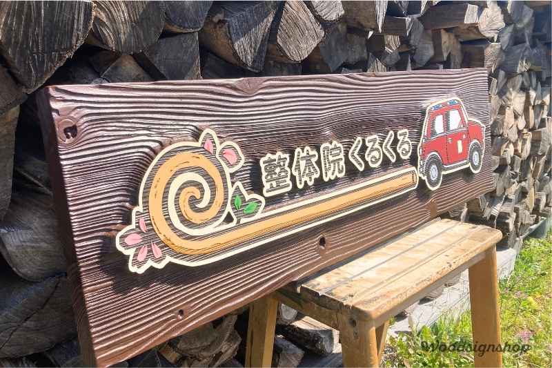 木製看板を左側から斜めに撮影した写真。「整体院くるくる」の文字と渦巻きのロゴマークがぷっくりと浮き出るように彫刻されており、背景の木目が深く美しい波模様を描いている。