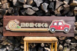 背景に薪が積まれた、木製の椅子の上に載せられた「整体院くるくる」の木製看板正面全体。左側に渦巻きと桜のロゴ、中央に店名、右側に赤いローバーミニのイラストが立体的に彫刻されている。