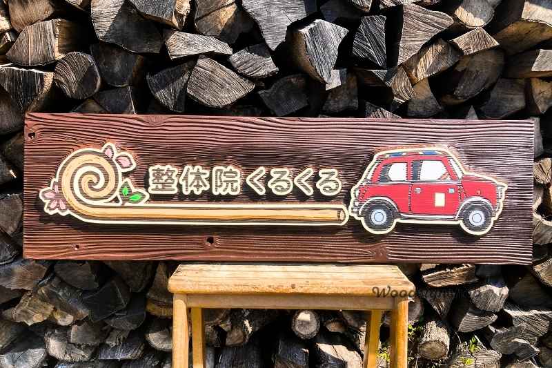背景に薪が積まれた、木製の椅子の上に載せられた「整体院くるくる」の木製看板正面全体。左側に渦巻きと桜のロゴ、中央に店名、右側に赤いローバーミニのイラストが立体的に彫刻されている。