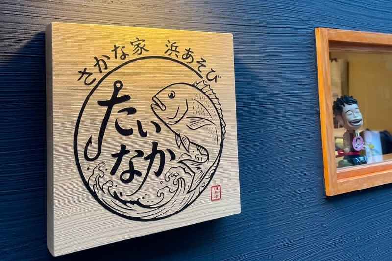 シックな外壁に取り付けられた「さかな家 浜あそび たいなか」様の壁掛け木製看板