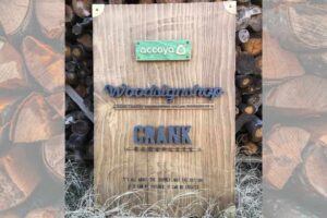 薪を背景に立てかけた、accoya®︎の木材で作った展示用看板。上から緑のaccoyaプレート、Woodsignshopの切り文字、CRANK NAMEPLATEの切り文字が並ぶ
