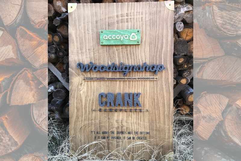 薪を背景に立てかけた、accoya®︎の木材で作った展示用看板。上から緑のaccoyaプレート、Woodsignshopの切り文字、CRANK NAMEPLATEの切り文字が並ぶ